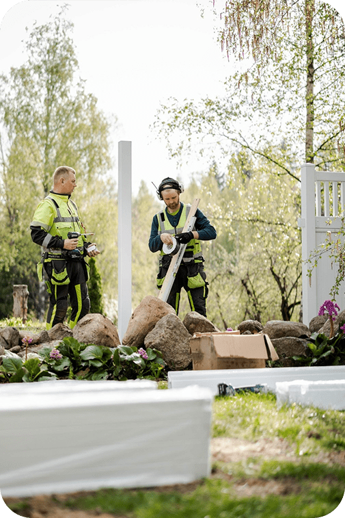 Kaksi työmiestä asentaa vinyyliaidan perustuksia omakotitalon pihalle